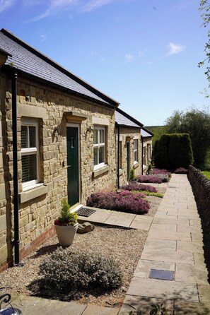 A row of stone buildings with a paved walkway, a green door, and a well-maintained garden with purple flowers. - 1 The Old Stables is a rural cottage with scenic countryside views. (Knitsley)