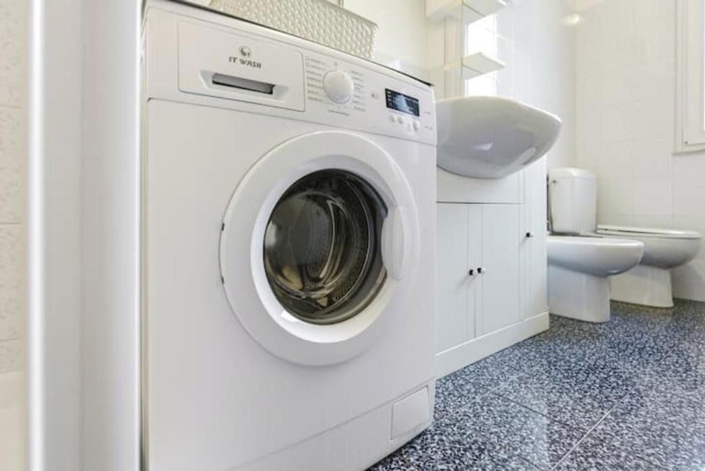 A front-loading washing machine in a bathroom with a toilet and a sink.