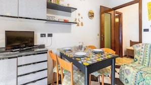 Een compacte woonruimte met een eettafel voor vier personen, een bank, een televisie, en een keuken met kastjes en een magnetron.