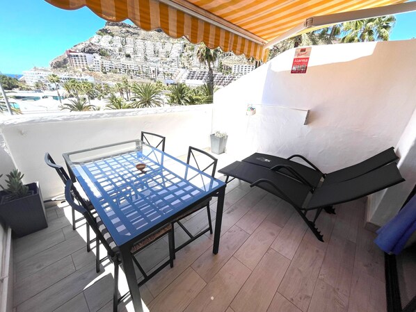 A balcony with a table and chairs, a sun umbrella, and a view of a hillside with buildings.