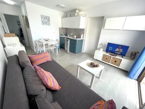 Ein modernes Wohnzimmer mit einem grauen Sofa, einem kleinen weißen Tisch, einem Fernseher und einer Küchenzeile mit Schränken und einem Esstisch.