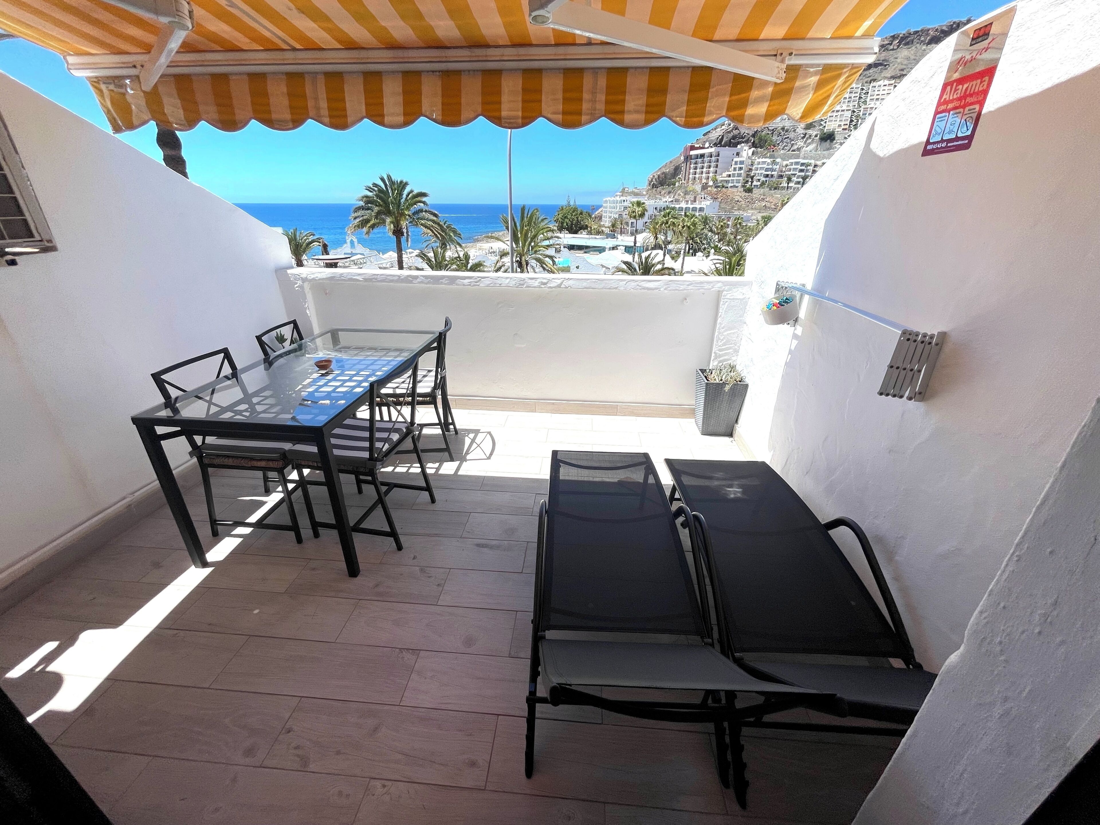 A balcony with a table and chairs, a sun lounger, and a view of the sea and buildings.
