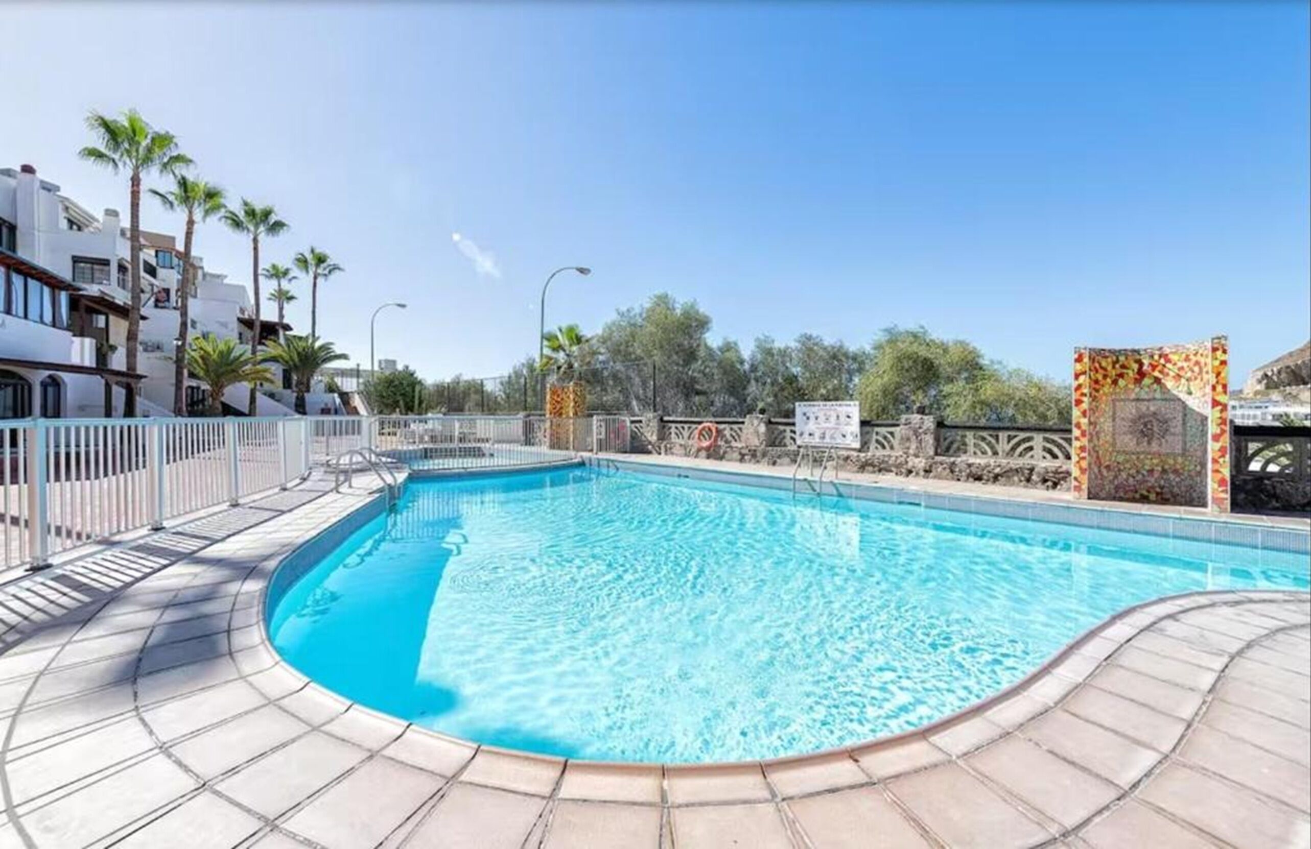 A swimming pool with a tiled deck, palm trees, and a clear sky.