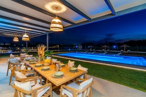 Ein Essbereich im Freien mit Pool, Holzmöbeln und hängenden Lampen.