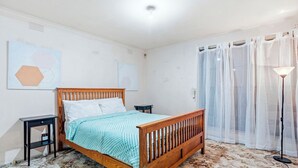 A bedroom with a wooden bed, a nightstand, a lamp, and a window with sheer curtains. - Well-Located St Albans Stay with Fully Equipped Kitchen (Saint Albans)