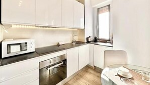 Una cocina moderna con armarios blancos, microondas, horno, pileta y un comedor con mesa de vidrio.
