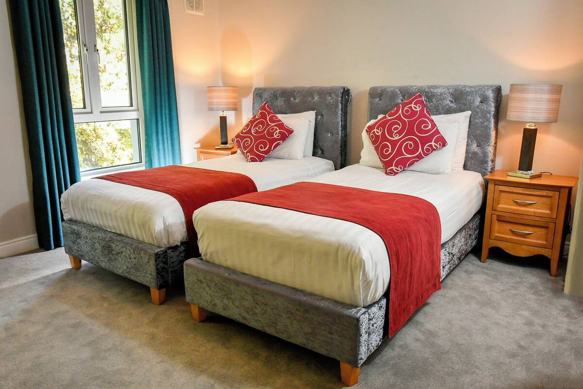 A double bed with a grey headboard, two bedside tables with lamps, and a window with curtains.
