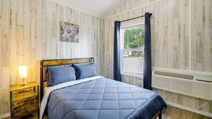 Un dormitorio con una cama, una mesita de noche, un ventanal con cortinas, un calentador en la pared y un cuadro con motivos florales.