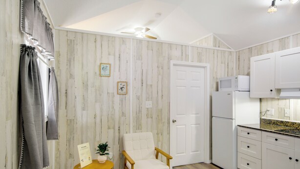 Una cocina compacta con armarios blancos, microondas y refrigerador. Hay una silla de madera y una mesa pequeña con una planta.