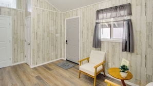 Una habitación con piso de madera, una puerta blanca, una silla blanca, una mesita redonda con una planta y una ventana con cortinas.