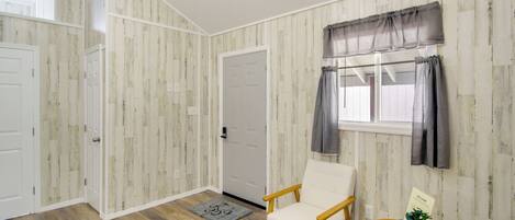 A room with a wooden floor, a white door, a white chair, a small round table with a plant, and a window with curtains.