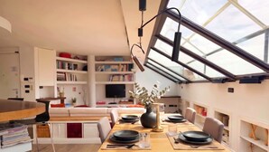Et moderne spiseområde med et træbord dækket til fire personer, en bogreol i baggrunden og et ovenlysvindue.
