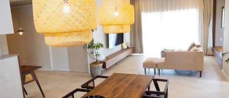 A modern living room with a wooden dining table, two hanging rattan lamps, a sofa, and a TV mounted on the wall.