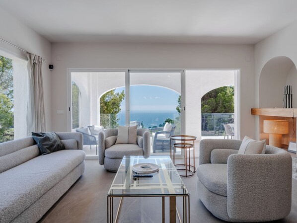 Ein modernes Wohnzimmer mit einem Glastisch, Sofas, Sesseln, Meerblick durch große Fenster und einem Kamin mit einer Lampe.