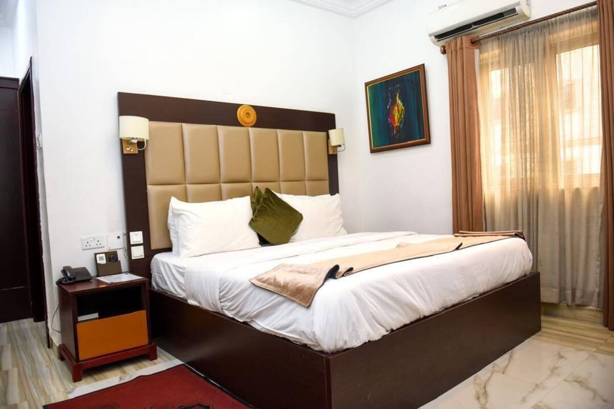 A hotel room with a large bed, a nightstand, a wall-mounted lamp, a painting, and a window with curtains.