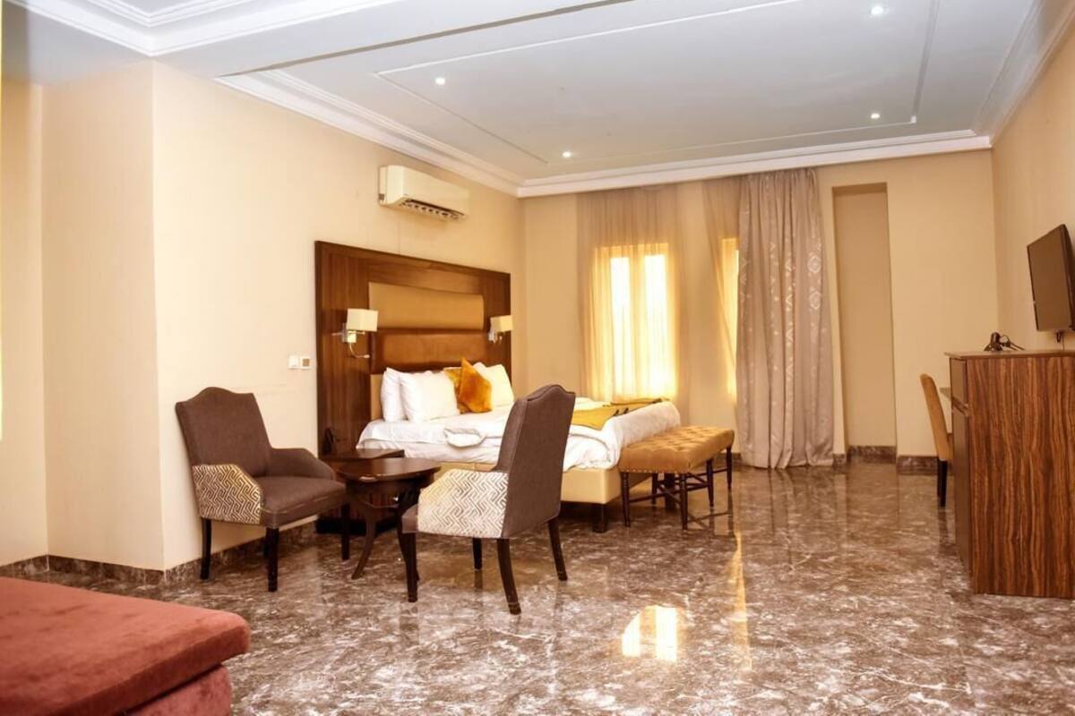 A hotel room with a bed, two armchairs, a desk, a TV, and a marble floor.