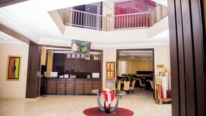 Vestíbulo de hotel con mostrador de recepción, zona de comedor, escalera y televisor en la pared.