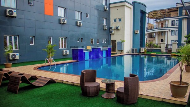 Una piscina con sillones de descanso y césped artificial, rodeada por un edificio con varias unidades de aire acondicionado.