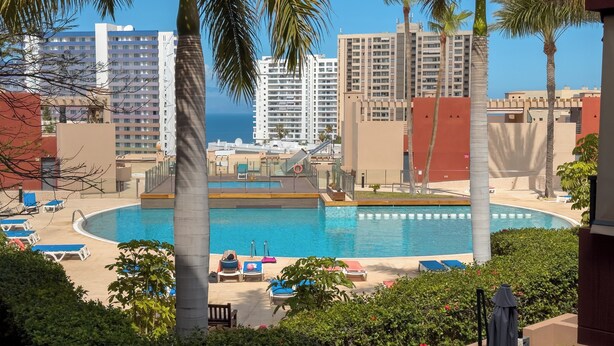 A pool area with lounge chairs, palm trees, and high-rise buildings in the background. - Duplex with sea view (Adeje)