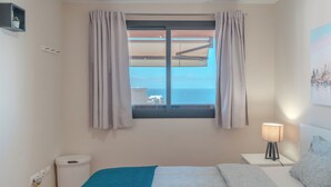 A bedroom with a bed, a nightstand with a lamp, a window with curtains, a view of the sea, and a small shelf with a plant. - Duplex with sea view (Adeje)
