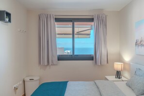 A bedroom with a bed, a nightstand with a lamp, a window with curtains, a view of the sea, and a small shelf with a plant. - Duplex with sea view (Adeje)