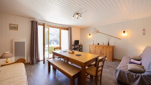 A room with a wooden table, bench, and a sofa. There is a television on a stand, a cabinet, and a window with curtains.