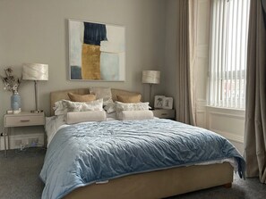A bedroom with a bed, two bedside tables, a lamp, a painting, and a window with curtains.