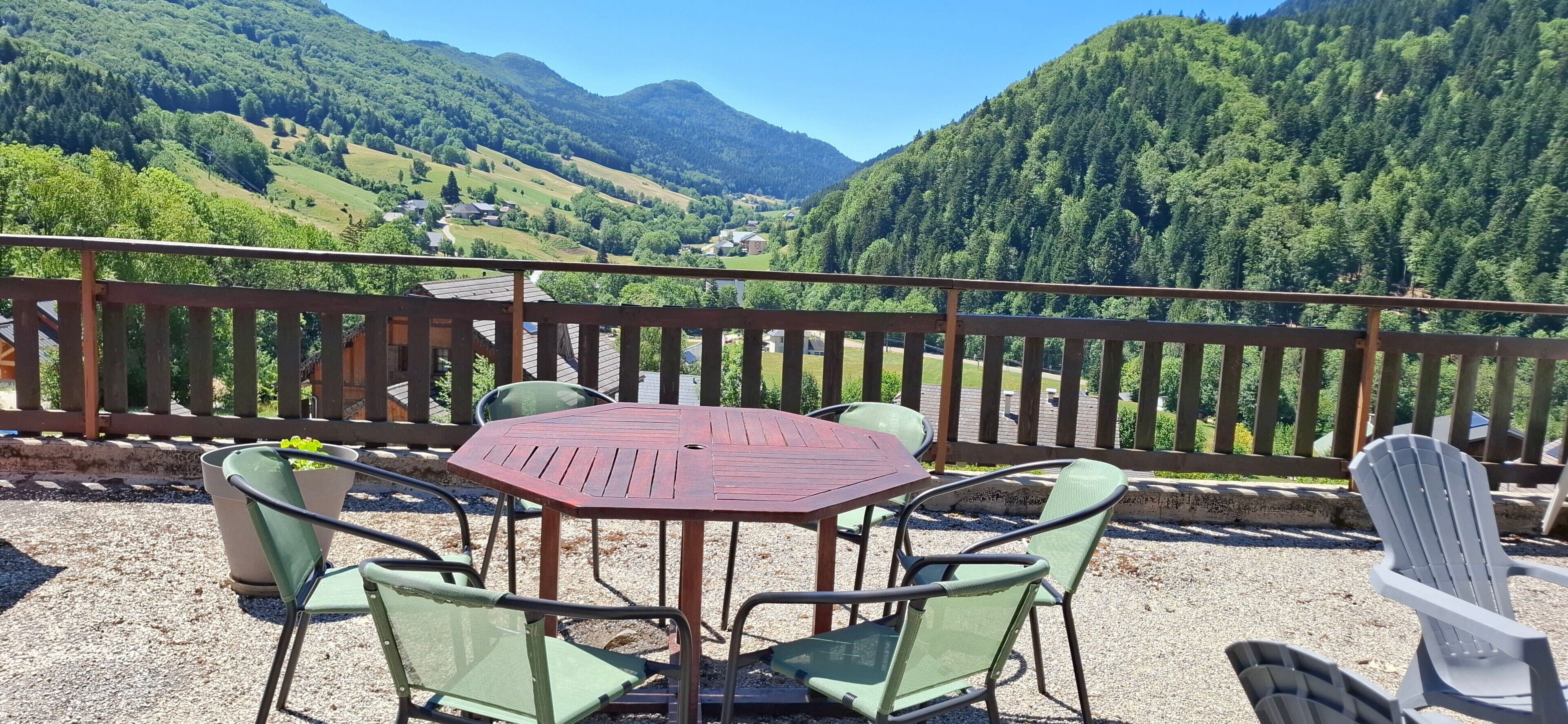 A terrace with a table and chairs, a wooden railing, and a view of a mountainous landscape.