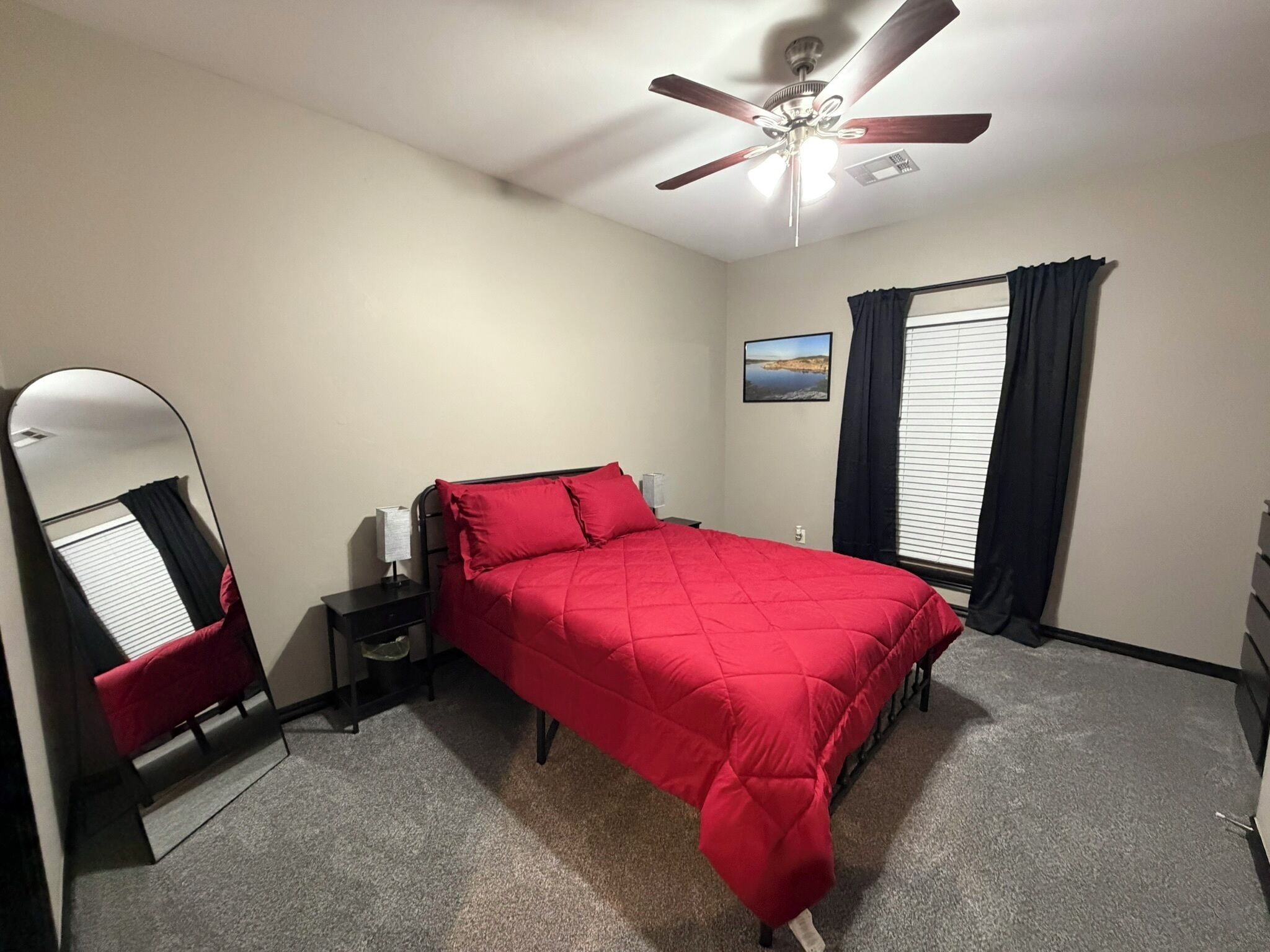 Um quarto com cama, mesa de cabeceira, espelho, ventilador de teto, janela com cortinas e uma pequena pintura na parede.