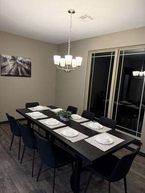 A dining room with a long table set for six, black chairs, a chandelier, and a painting on the wall.