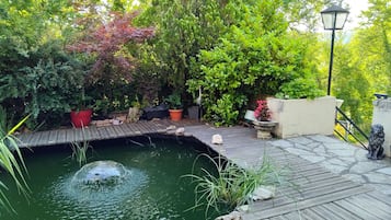 Une terrasse en bois avec un petit étang, entourée de différentes plantes en pot et d’un mur de pierre.