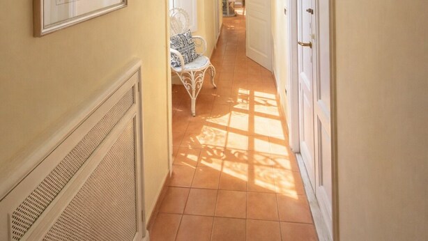 Un couloir avec un sol carrelé, un tableau encadré accroché au mur, une chaise blanche et une porte donnant sur une autre pièce.