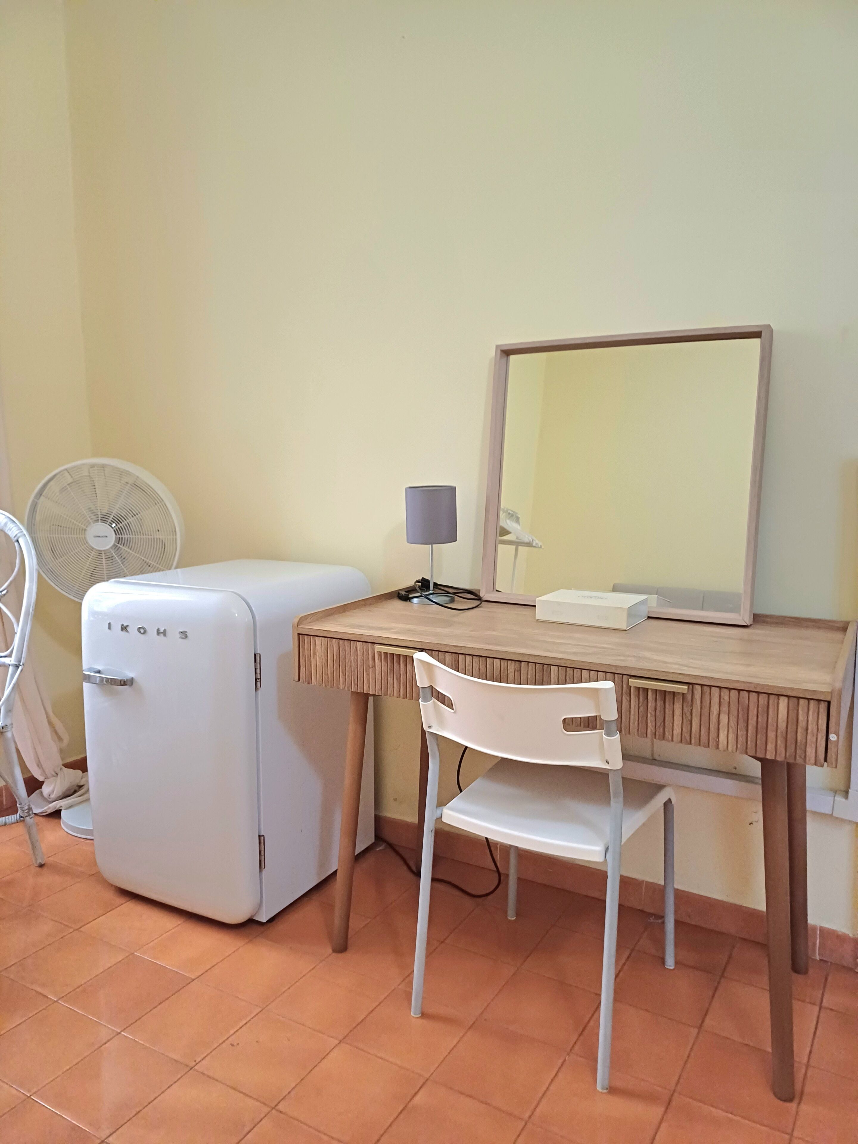 A compact room with a wooden desk, a white chair, a small white refrigerator, a standing fan, and a mirror on the wall.