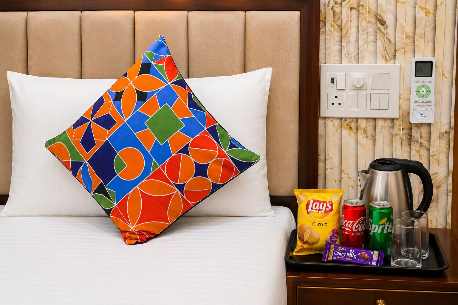Une chambre d’hôtel avec un lit, un oreiller coloré, un plateau avec des en-cas, une bouilloire et une prise électrique.