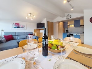 Un salon moderne avec un canapé, une table à manger dressée avec des assiettes, des verres à vin et une bouteille. On aperçoit, au fond, une cuisine avec des placards bleus et une horloge.