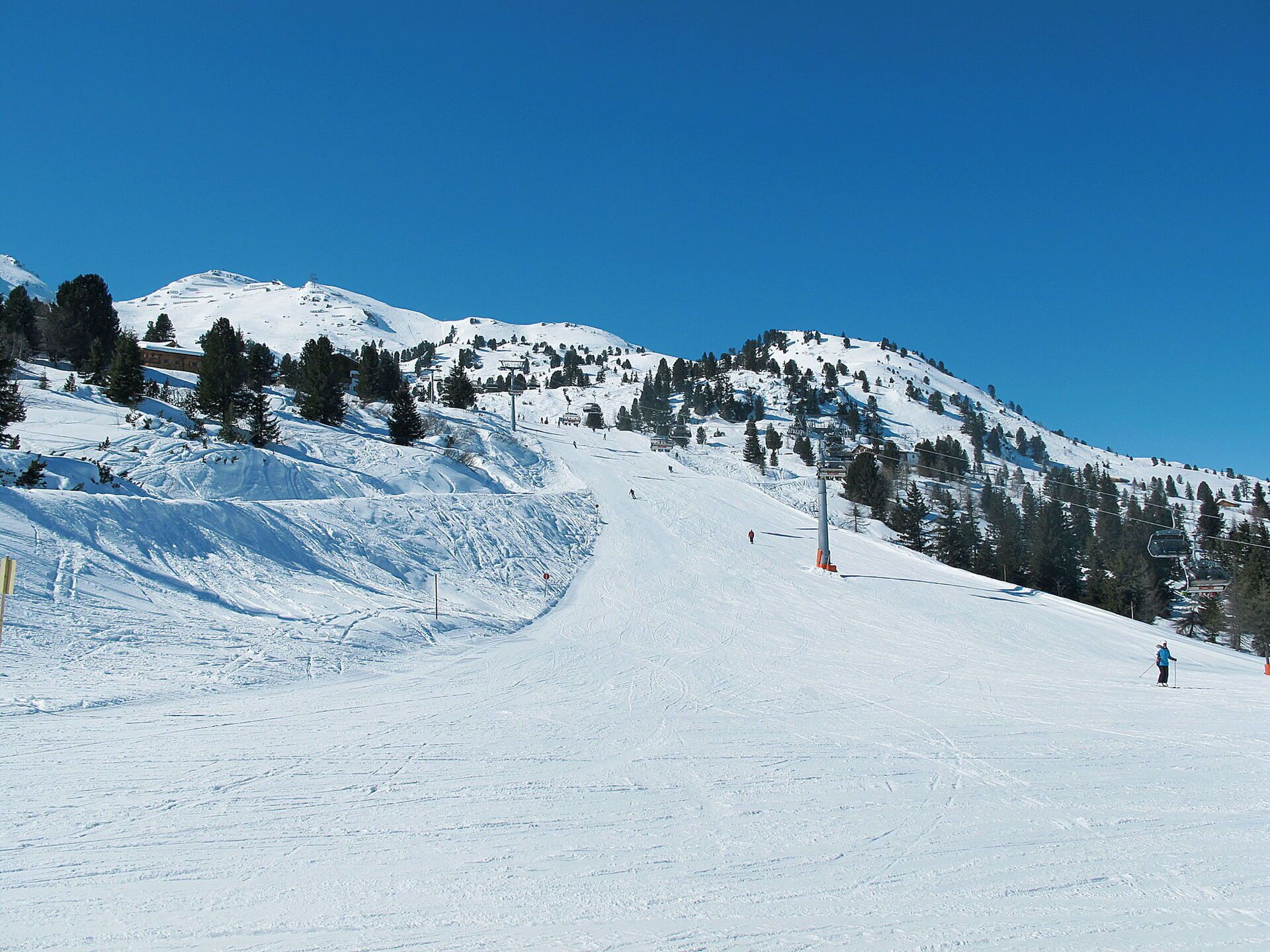 Pista da sci innevata con cielo azzurro, alberi e alcuni sciatori.