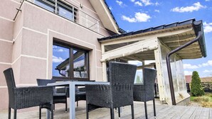 Un patio al aire libre con mesa y sillas, una zona cubierta y un edificio con ventanas amplias.