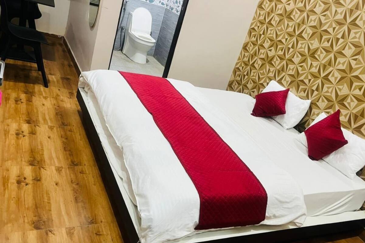 Ein Bett mit roter und weißer Bettdecke, Holzboden, ein schwarzer Stuhl und ein Badezimmer mit weißer Toilette und Spiegel.