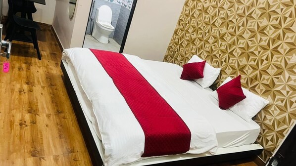 Una cama con edredón rojo y blanco, suelo de madera, una silla negra y un baño con inodoro blanco y espejo.