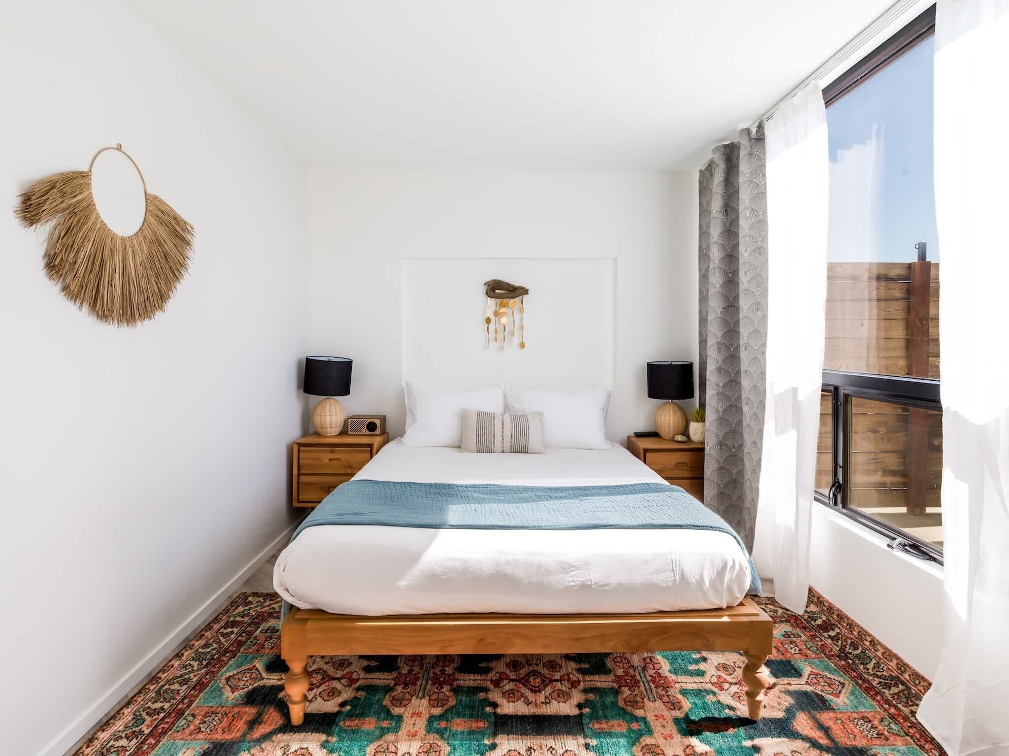 A bedroom with a bed, two bedside tables, a wall hanging, and a large window.
