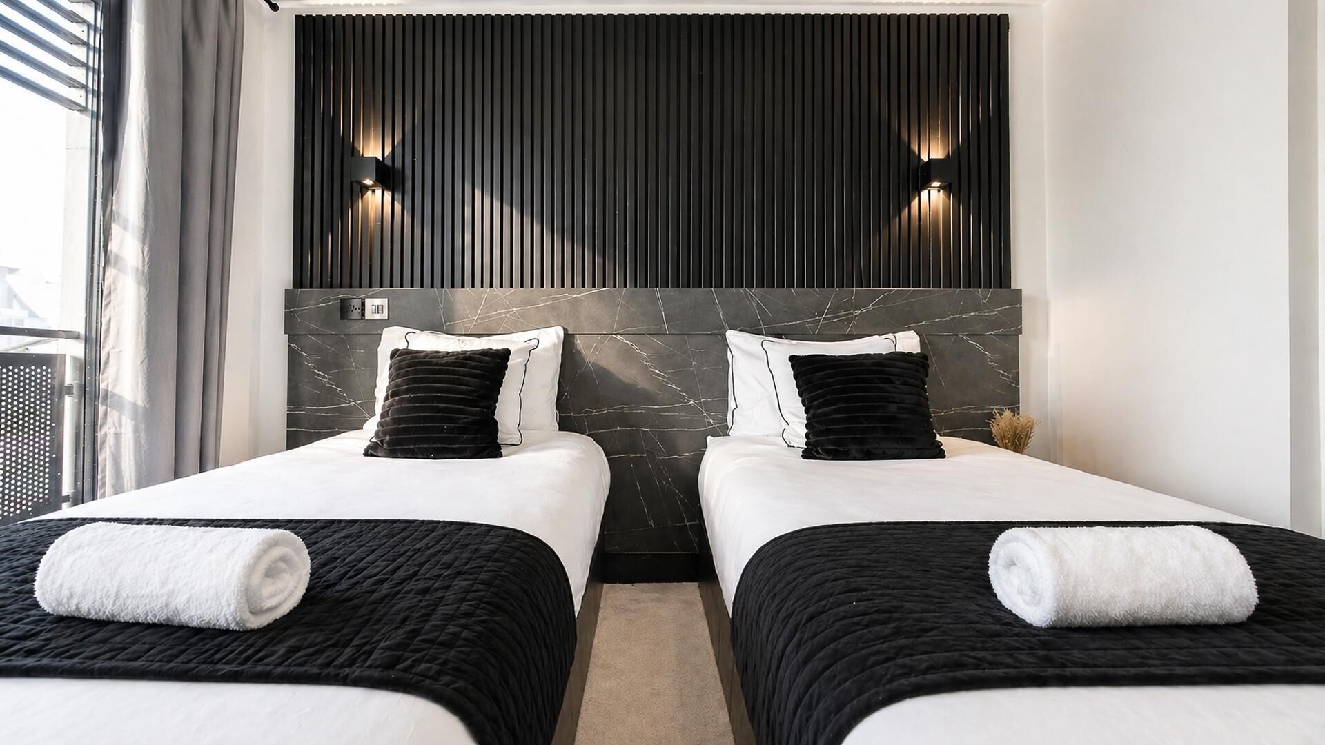 Ett hotellrum med två sängar, en mörk sänggavel och väggmonterade lampor.