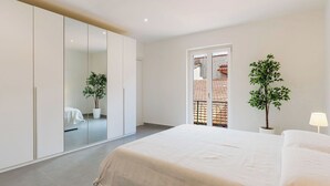 Une chambre avec un grand lit, une armoire, un balcon avec une balustrade et deux plantes en pot.