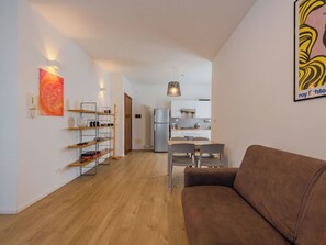 A modern living space with a brown sofa, a dining area with a table and chairs, a kitchen with white cabinets, and a hallway leading to a door. - Lovely apartment for 4 people with WIFI, A/C, TV, patio and panoramic view (Arco)