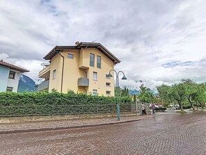 Ein mehrstöckiges Gebäude mit Balkon, gepflasterter Straße und Bergelandschaft im Hintergrund.