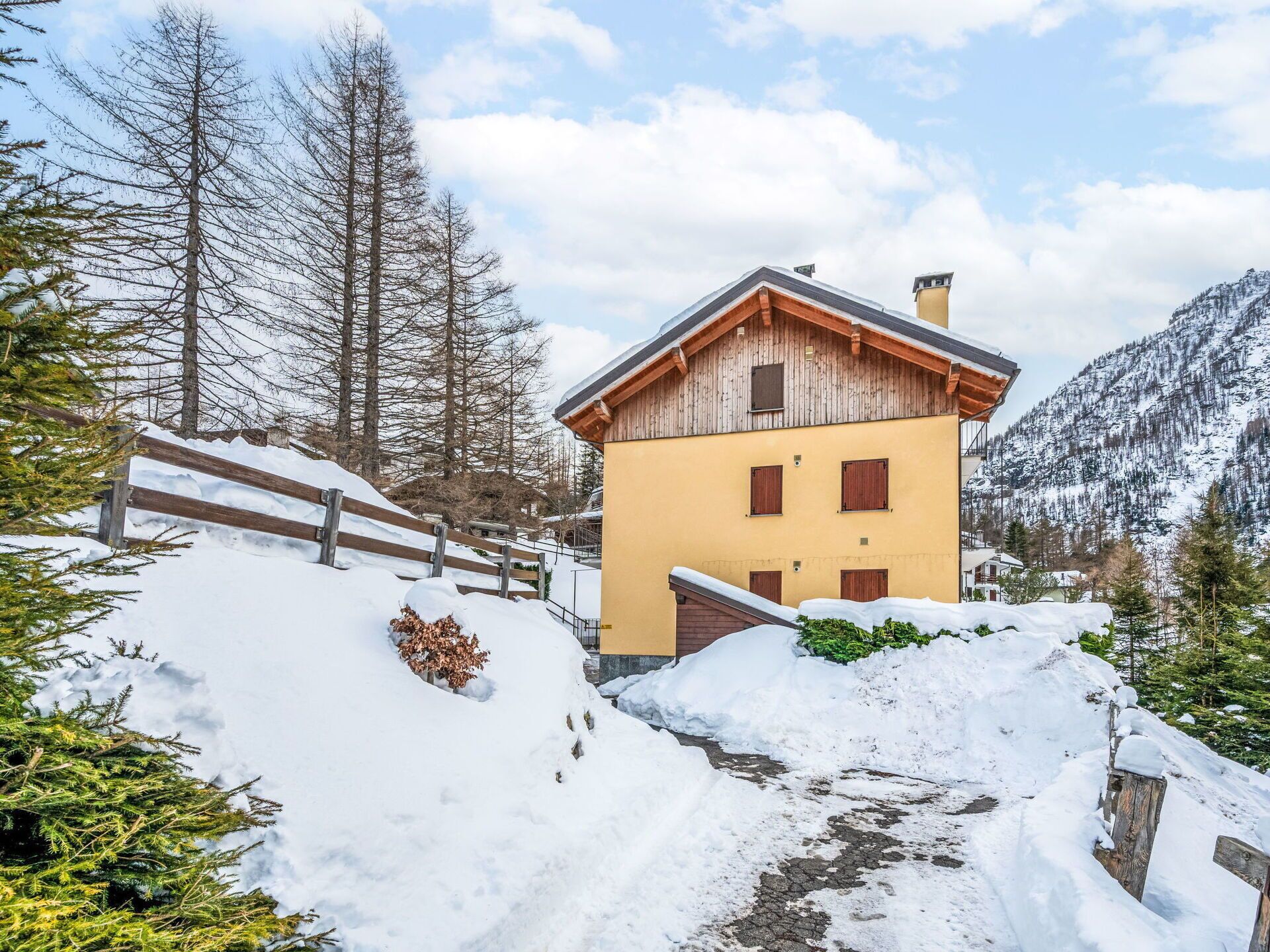 Un edificio amarillo con contraventanas de madera, rodeado de terreno y árboles cubiertos de nieve, con una montaña al fondo.
