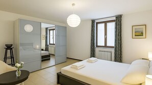 Una habitación de hotel con cama, mesita de noche, lámpara, silla, una mesa redonda, un jarrón con flores y un espejo grande.