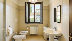 Un baño con inodoro, bidé, lavamanos y una ventana con contraventanas abiertas.