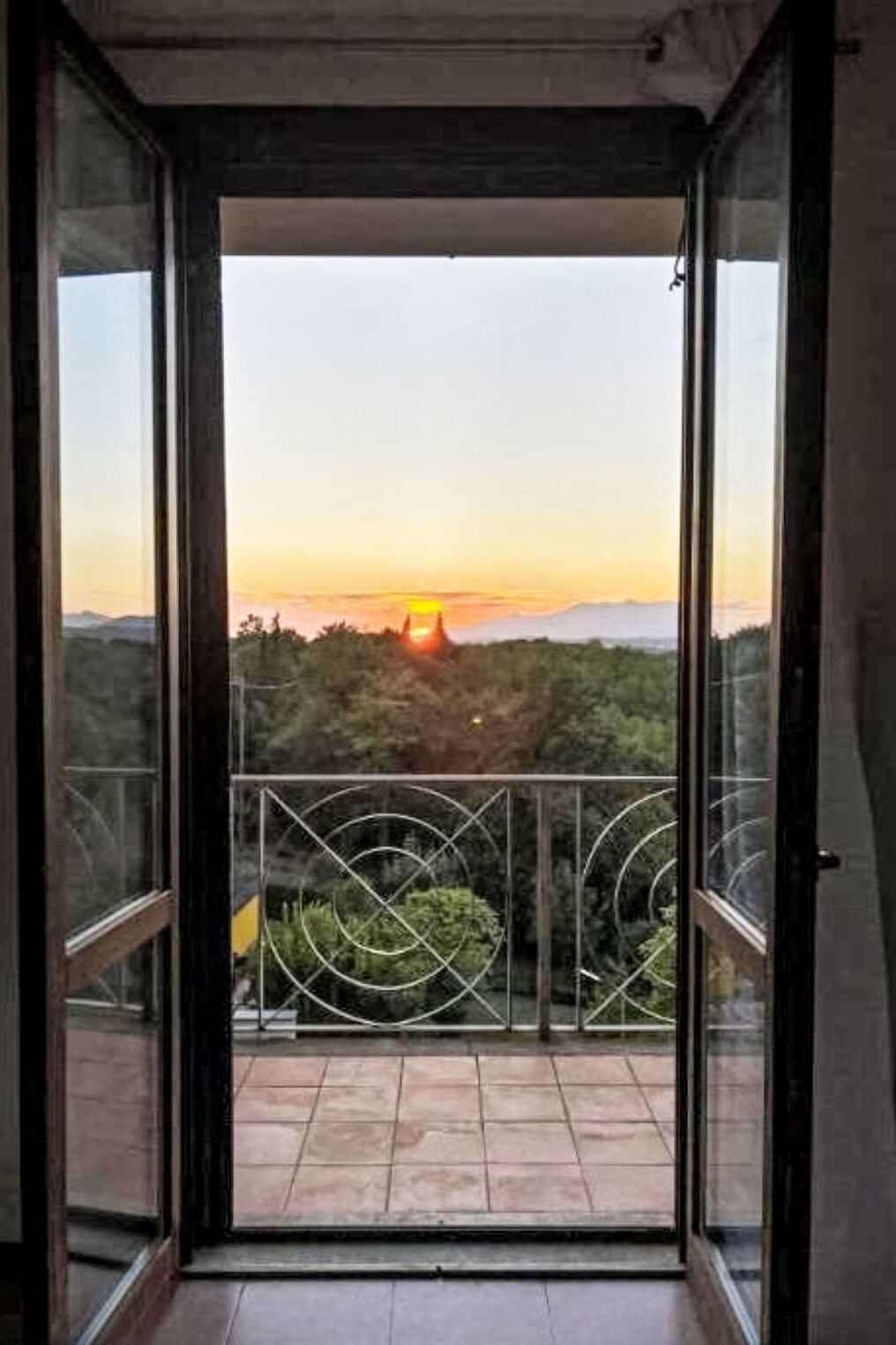 Puerta abierta que da a un balcón con vista al atardecer sobre un paisaje montañoso.