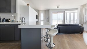 Une cuisine moderne dotée d’un îlot central, de tabourets de bar, d’un canapé de couleur sombre et de grandes fenêtres offrant une vue dégagée.