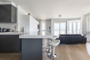 A modern kitchen with a central island, bar stools, a dark-colored sofa, and large windows offering a view. - Malibu Beach 0201 + P* (Sint Idesbald)